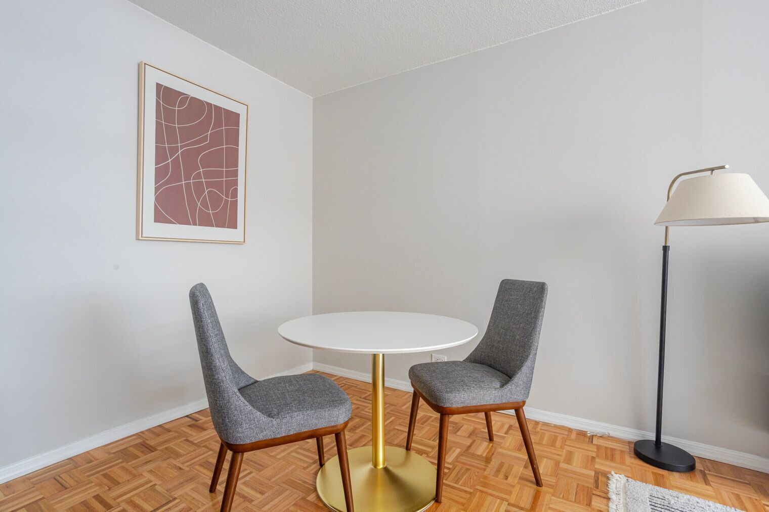 Una habitación con una mesa redonda, dos sillones grises tapizados, una base dorada, una lámpara de pie y una obra de arte abstracto enmarcada en la pared.