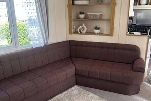 Un salon d’angle avec un canapé marron foncé, une étagère en bois ornée d’objets décoratifs et un coin cuisine équipé d’une cuisinière.