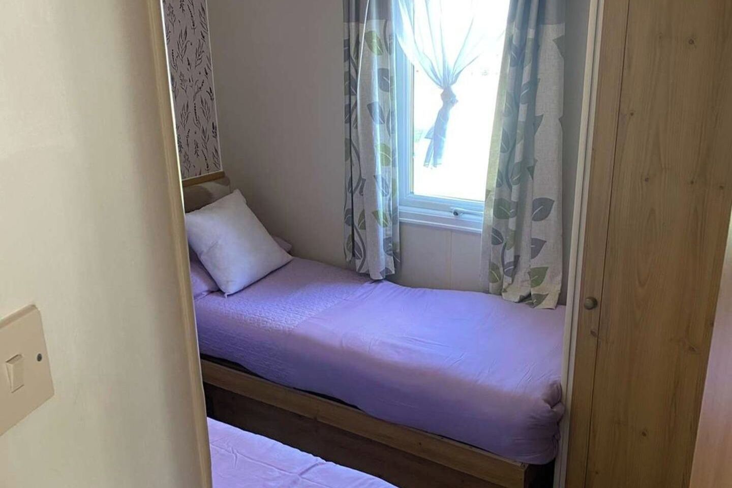 Un lit simple avec un cadre en bois et un matelas blanc, une fenêtre avec des rideaux et un mur recouvert d’un papier peint à motifs de feuilles.
