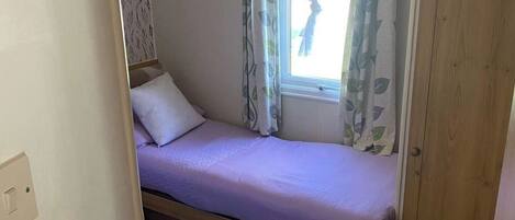 Un lit simple avec un cadre en bois et un matelas blanc, une fenêtre avec des rideaux et un mur recouvert d’un papier peint à motifs de feuilles.