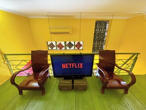Ein Zimmer mit einem Fernseher, der 'NETFLIX' anzeigt, zwei Holzstühlen, einem grünen Boden und einer gelben Wand mit einem Zierfenster.