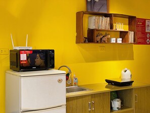 A compact kitchen with a white refrigerator, a microwave on top, a sink, and a shelf with books and kitchenware.
