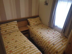 Ein kleines, gemütliches Zimmer mit zwei Einzelbetten, einem Holzkopfende, einem kleinen Tisch und einem Fenster mit Vorhängen.
