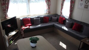 Ein Wohnzimmer mit einem grauen Ecksofa, einem Flachbildfernseher, einem Couchtisch mit Pflanze und einem fenster mit Vorhängen.