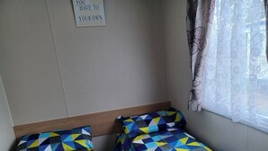 Ein kleines Zimmer mit zwei Einzelbetten, einem hölzernen Kopfteil, einem an der Wand befestigten Schild und einem Fenster mit durchsichtigen Vorhängen.