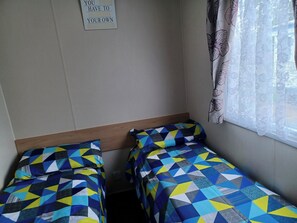 Ein kleines Zimmer mit zwei Einzelbetten, einem hölzernen Kopfteil, einem an der Wand befestigten Schild und einem Fenster mit durchsichtigen Vorhängen.