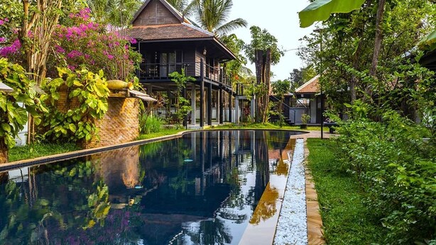 A pool surrounded by lush greenery and a traditional house with a balcony.
