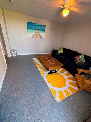 Ein Wohnzimmer mit einem dunkelgrauen Sofa, einem Holztisch, einem Teppich mit Sonnendesign, einem Deckenventilator und einem an der Wand hängenden Bild von einem Strand.