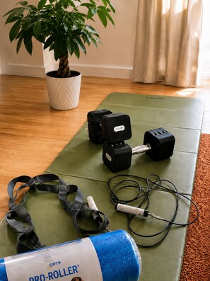 A yoga mat, two dumbbells, resistance bands, a jump rope, and a blue yoga mat on a wooden floor with a potted plant and a curtain in the background.