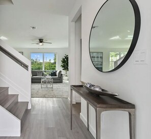 A modern interior with a staircase, a round mirror, and a view to a living area with a sofa and plants. - Modern Luxury 4BR Oasis - Near Beaches, Airport & Downtown (West Palm Beach)