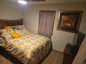 Ein Schlafzimmer mit einem Bett, einem Fenster mit Vorhängen, einem gerahmten Bild an der Wand und einem kleinen Tisch mit einer Lampe.