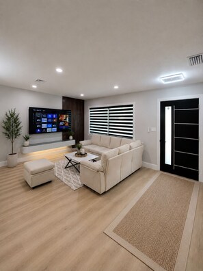 Ein modernes Wohnzimmer mit einem Ecksofa, einem Flachbildfernseher an der Wand, einem Topf mit Pflanze und einer schwarzen Tür mit vertikalem Griff.
