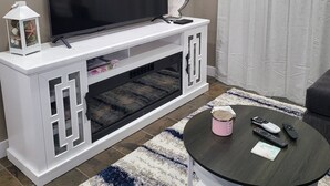 Ein Wohnzimmer mit einem Flachbildfernseher auf einer weißen Unterhaltungsanlage, einem Couchtisch mit Fernbedienungen und einem gemusterten Teppich.