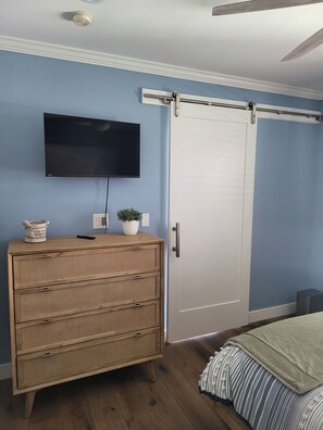 A bedroom with a wooden dresser, a flat-screen TV mounted on the wall, a sliding door, and a bed with striped bedding. - Tranquil 1-bedroom studio with AC in charming Chula Vista (Chula Vista)