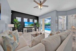 A living room with a flat-screen TV, a ceiling fan, and a large window overlooking a pool. - 5BR Disney Home, Private Pool, Spa & PS5 Game Loft (Davenport)