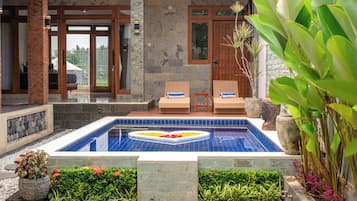 Ein Schwimmbecken mit einer herzförmigen Blumenkomposition, umgeben von einem Garten mit verschiedenen Pflanzen und einer Sitzgelegenheit mit zwei Korbstühlen.