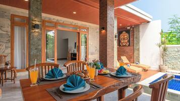 Un espace repas avec vue sur la piscine, comprenant une table dressée avec des assiettes, des serviettes et des boissons, ainsi qu’une statue de Bouddha accrochée au mur.