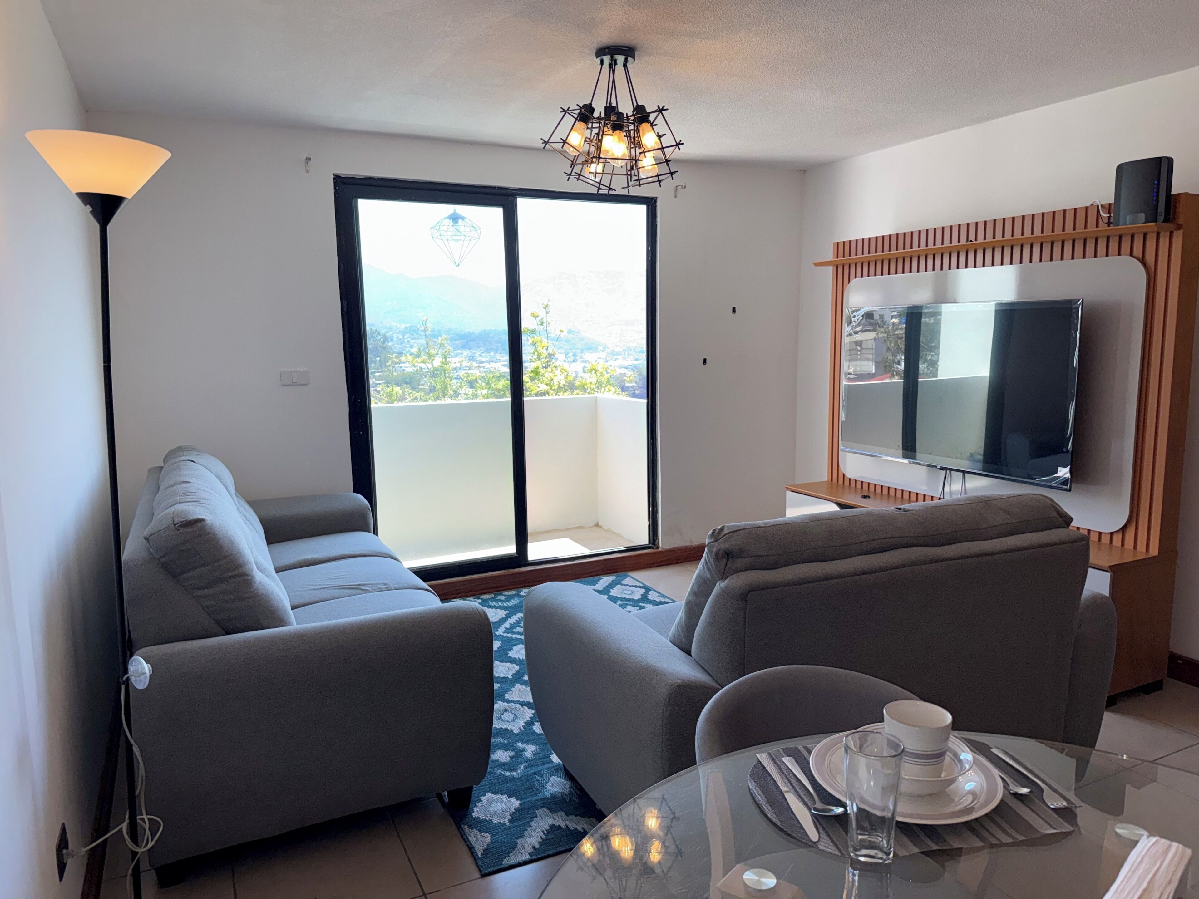 A modern living room with a sofa, a glass table set for dining, a television, and a view through a large window.