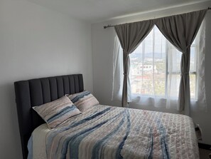 Een bed met een donker hoofdbord en strepenbeddengoed, een raam met gordijnen, en een uitzicht op gebouwen.