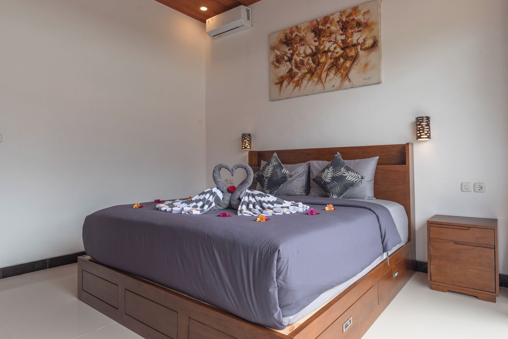 A neatly made bed with a heart-shaped pillow arrangement, a wall-mounted air conditioner, and a painting above the bed.