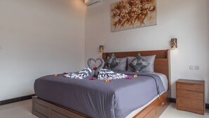 Uma cama bem arrumada com arranjo de almofadas em formato de coração, um ar condicionado fixado na parede e uma pintura sobre a cama.