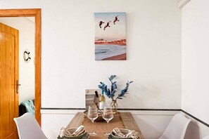 Ein Essbereich mit einem Holztisch, gedeckt für zwei Personen, einer Vase mit blauem Grünwerk und einem Gemälde von Vögeln über einem Strand.