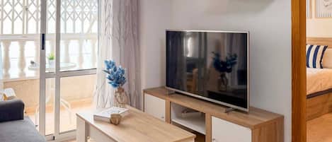En moderne stue med et fladskærms-TV på et træstel, en grå sofa, et lille hvidt bord med en bog og en kop, samt en dekorativ vase med blå blomster.