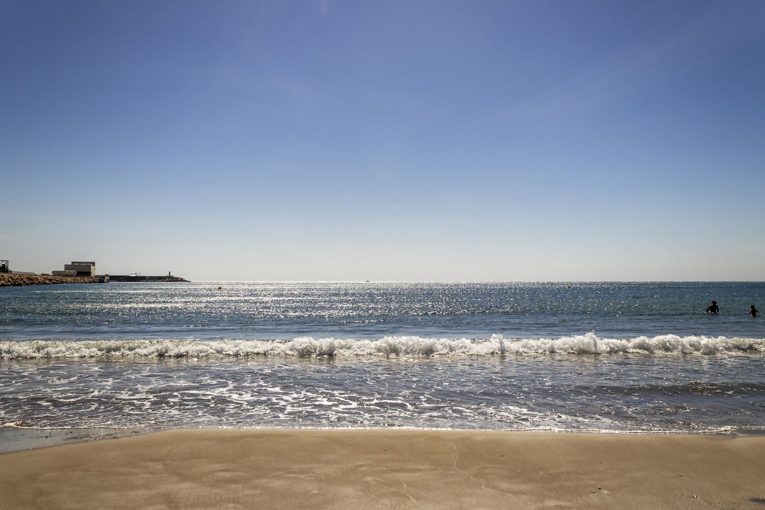 A beach with waves, a clear sky, and a distant shoreline with buildings.
