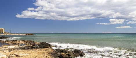 A rocky shoreline with waves, a clear sky, and distant buildings.