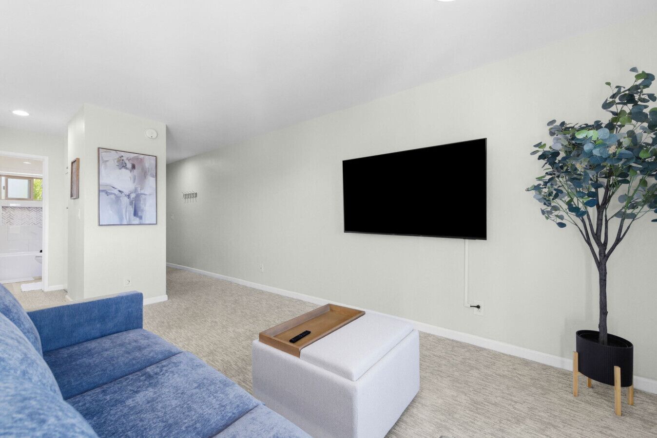 A modern living room with a flat-screen TV mounted on the wall, a blue sofa, a white ottoman, and a potted plant.