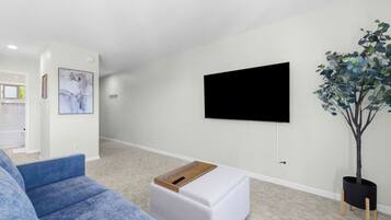 A modern living room with a flat-screen TV mounted on the wall, a blue sofa, a white ottoman, and a potted plant.