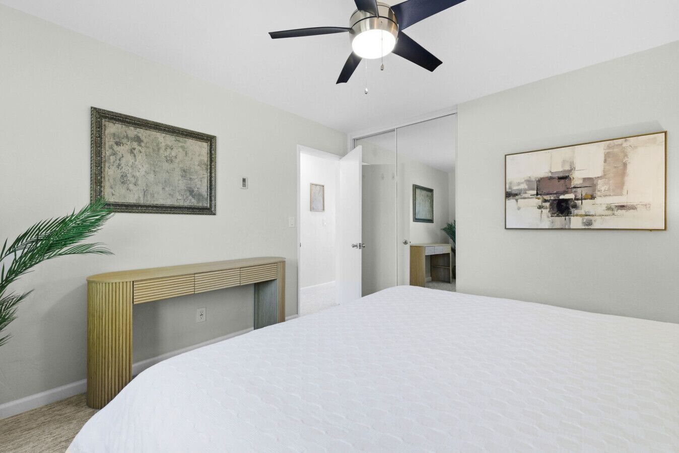 A bedroom with a bed, a ceiling fan, a console table, a painting, and a door leading to a bathroom.