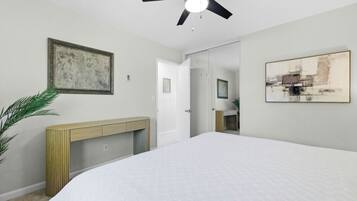 A bedroom with a bed, a ceiling fan, a console table, a painting, and a door leading to a bathroom.