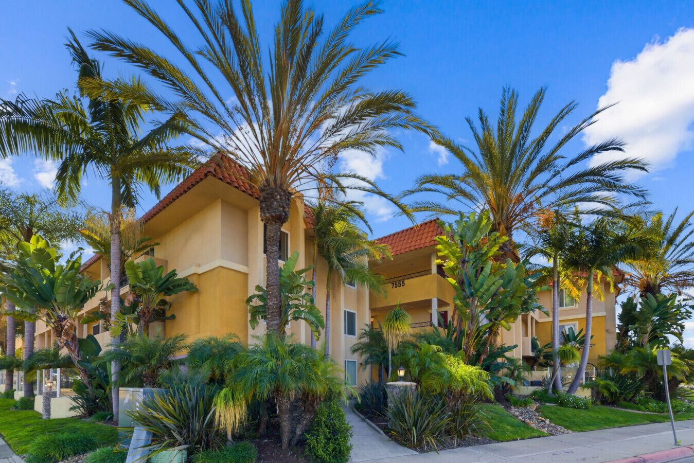 A multi-story hotel with a yellow facade and red-tiled roof, surrounded by palm trees and lush greenery.