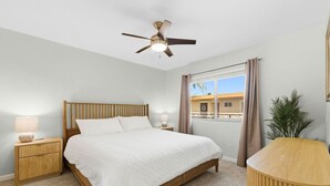 Quarto com cama, mesas de cabeceira, ventoinha no teto, janela com cortinas e vista para prédios.
