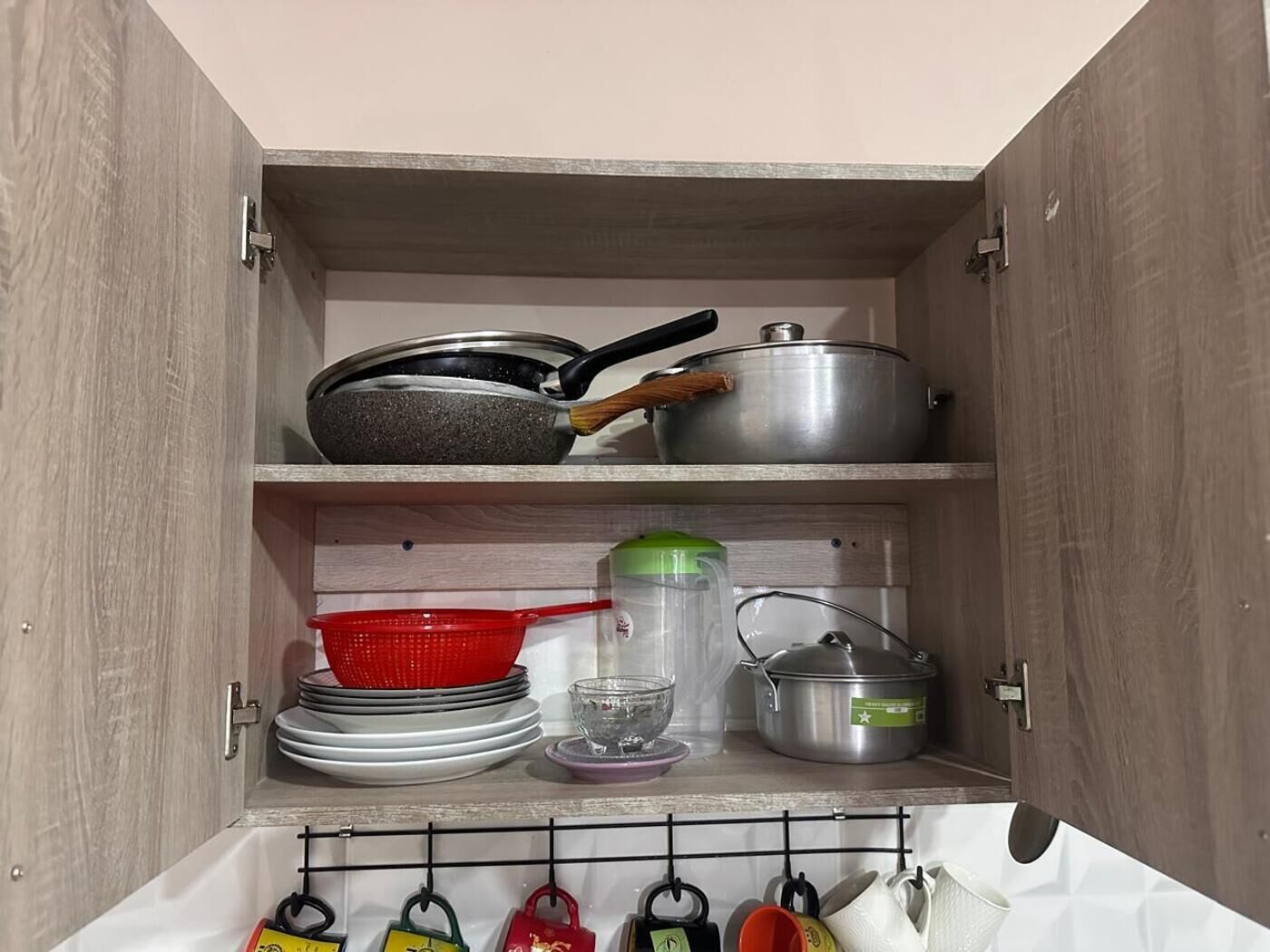A kitchen cabinet with cookware, a colander, a glass, and a hanging rack with utensils.