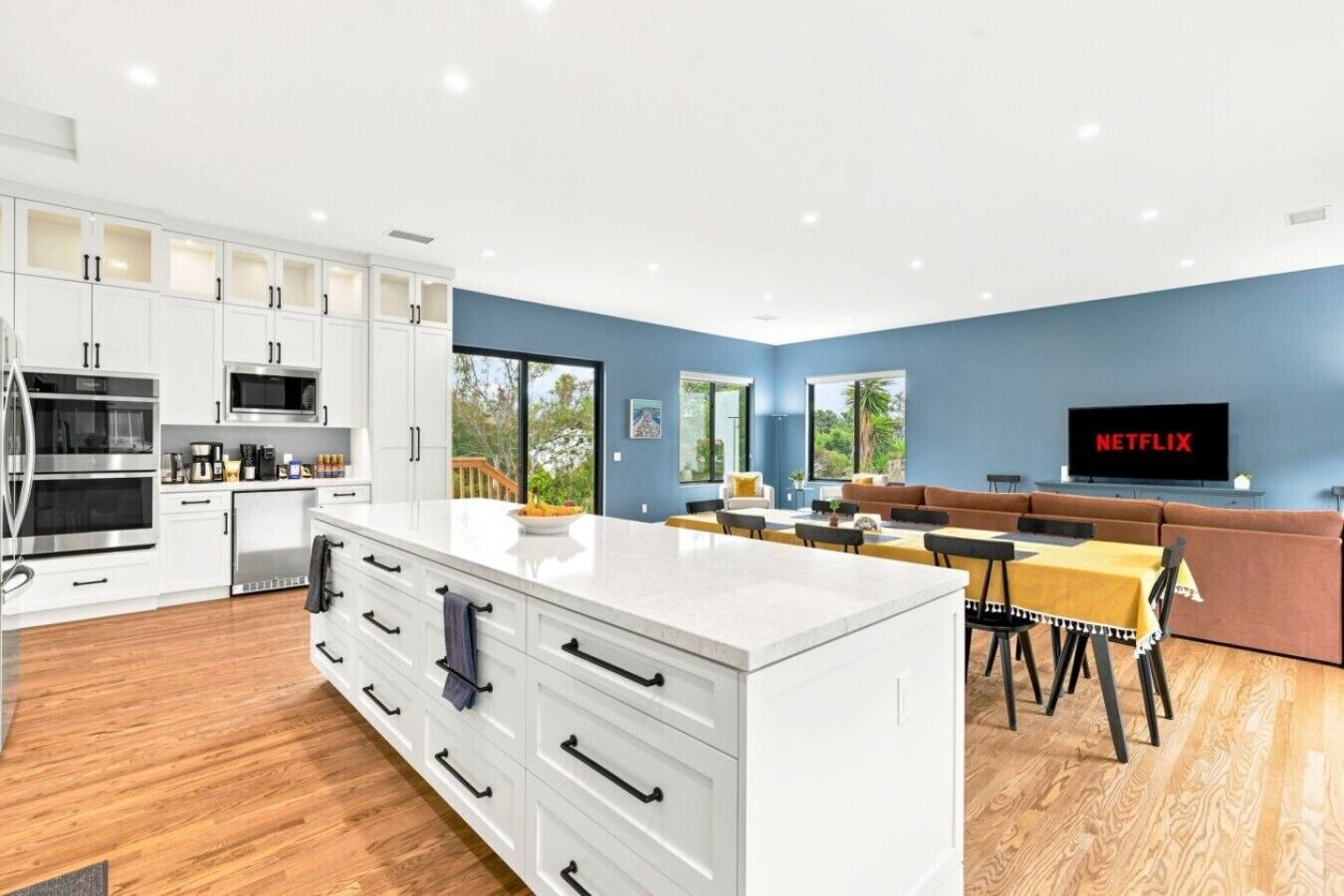 A modern kitchen with white cabinetry, a central island, and a dining area with a brown sofa and a TV displaying 'NETFLIX'.