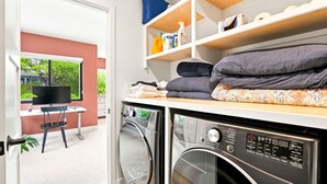 A laundry area with a washing machine, dryer, and shelves with folded clothes and laundry supplies. - Sunny Palms Luxury Home - Northpark (San Diego)