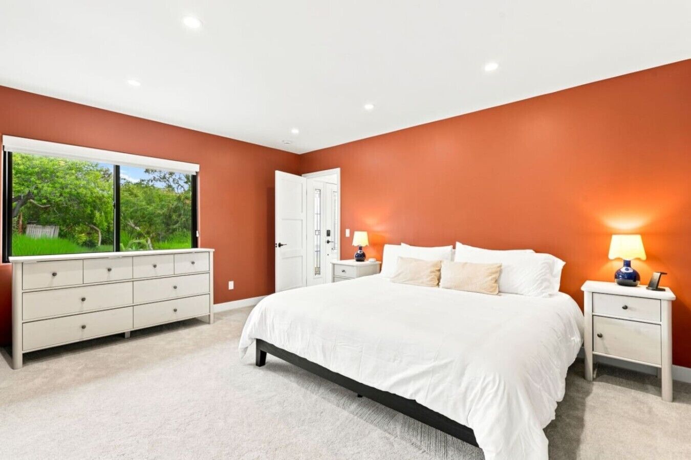 A bedroom with a large bed, a dresser, bedside tables, and a window with a view of greenery.