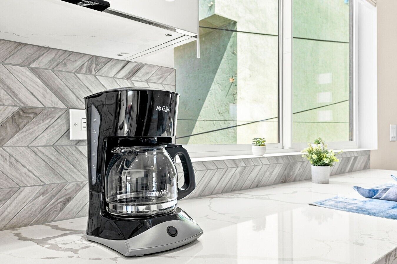 A modern coffee maker on a kitchen counter with a herringbone-patterned backsplash and a window with a view of greenery.
