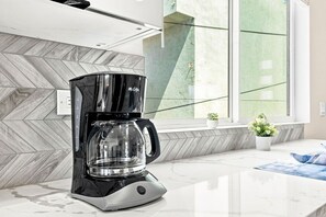 A modern coffee maker on a kitchen counter with a herringbone-patterned backsplash and a window with a view of greenery. - Brand New Condo Near Downtown, Coronado (San Diego)