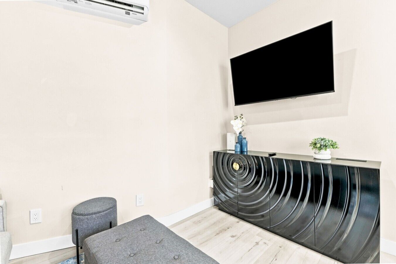 A modern living room with a flat-screen TV mounted on the wall, a black curved entertainment unit, a grey ottoman, and a small potted plant.