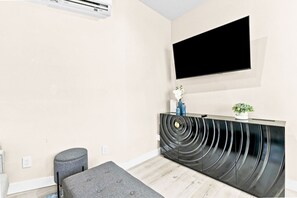 A modern living room with a flat-screen TV mounted on the wall, a black curved entertainment unit, a grey ottoman, and a small potted plant. - Brand New Condo Near Downtown, Coronado (San Diego)