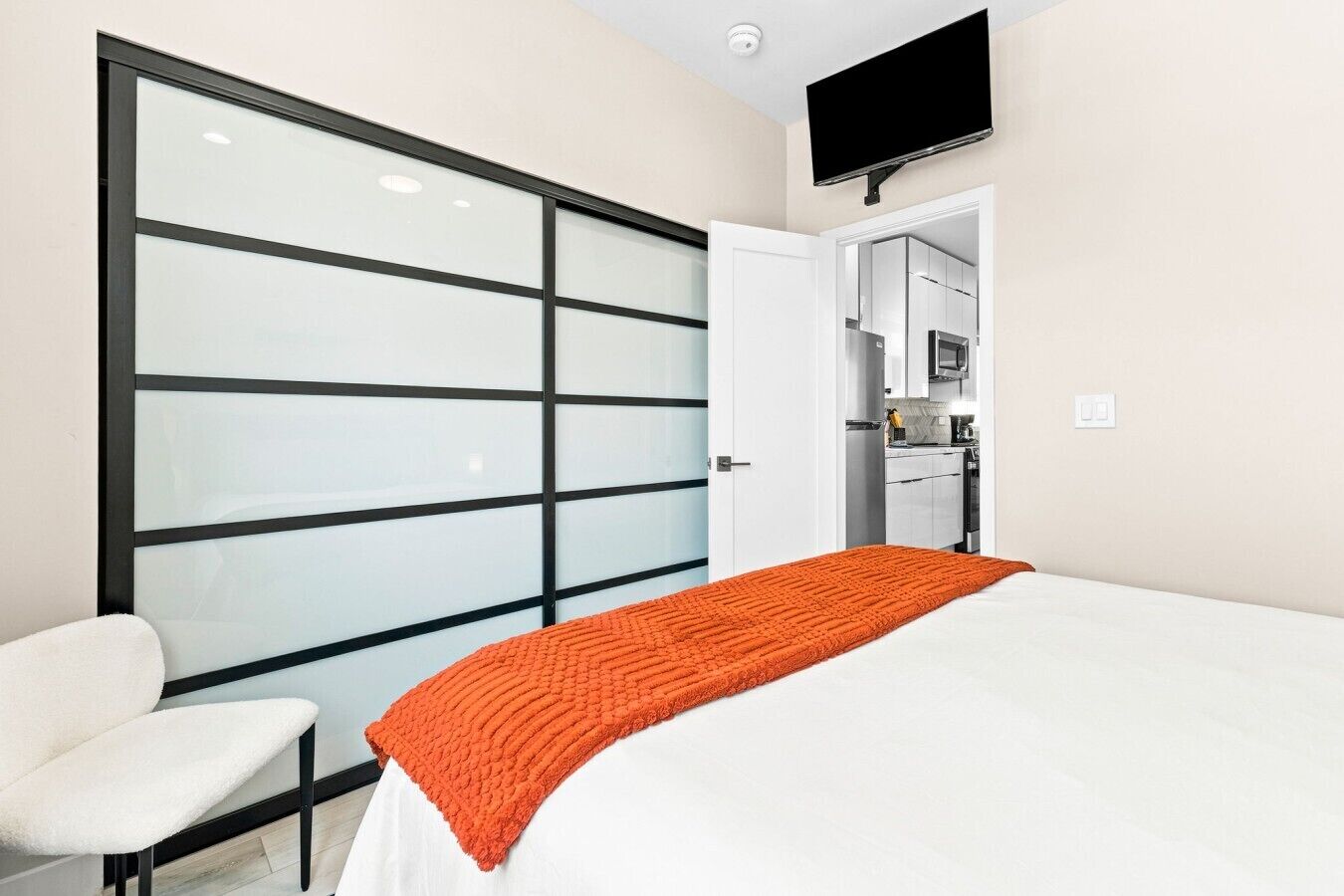 Ein Hotelzimmer mit einem Bett, einem weißen Sessel, einer Schiebetür, einem Fernseher und einer Küchenzeile.