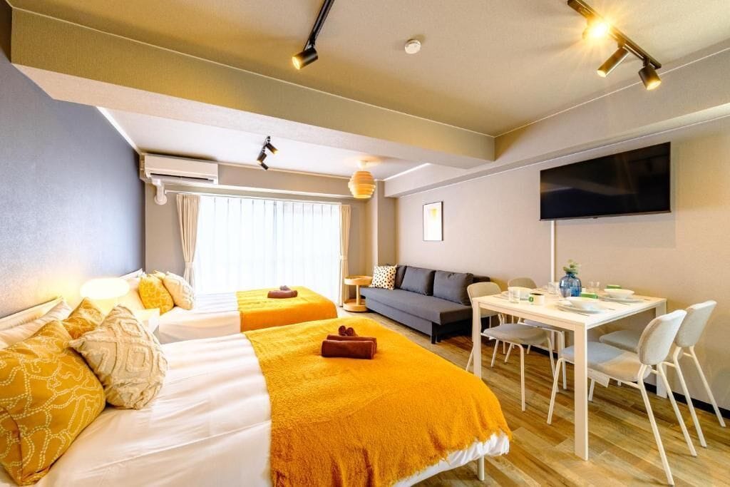 A hotel room with two beds, a sofa, a dining table with chairs, a flat-screen TV, and a window with curtains.