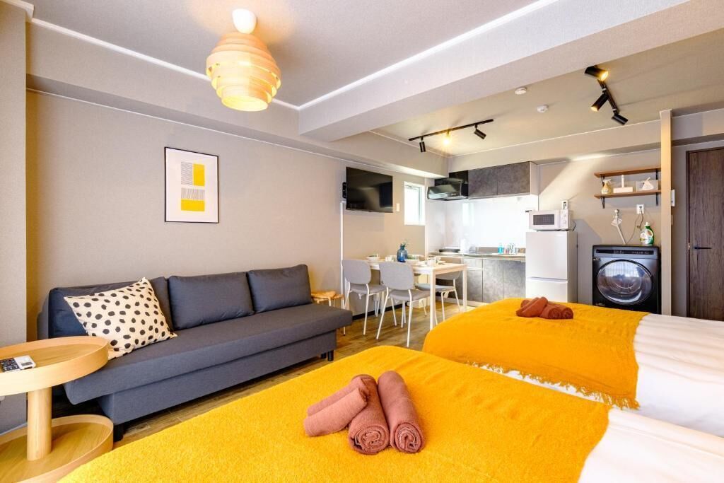 A modern hotel room with a sofa, two beds, a dining area, and a kitchen.