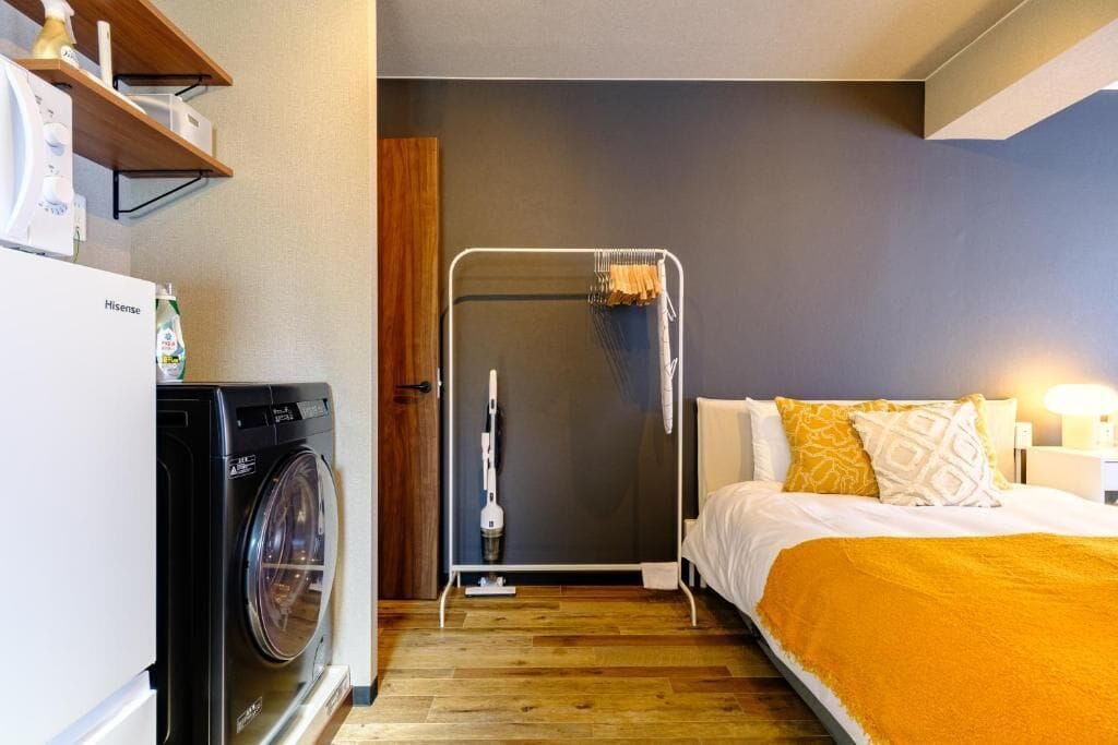 A compact hotel room with a washing machine, a shower area, and a bed with a yellow throw blanket.