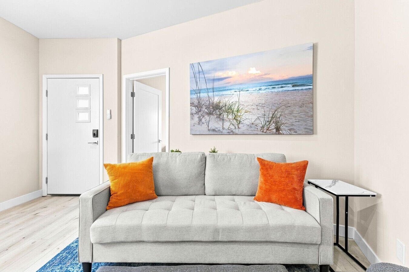 A living room with a grey sofa, two orange cushions, a small table, and a beach scene painting on the wall.
