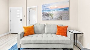 A living room with a grey sofa, two orange cushions, a small table, and a beach scene painting on the wall. - New 2BR Home Minutes to Downtown SD (San Diego)