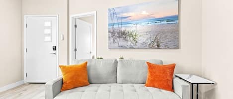 A living room with a grey sofa, two orange cushions, a small table, and a beach scene painting on the wall.